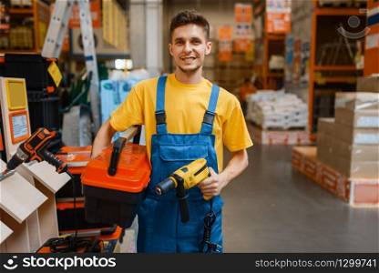 Male constructor holds tools in hardware store. Builder in uniform look at the goods in diy shop