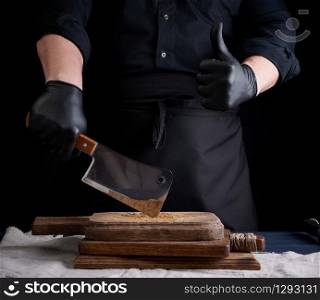 male cook in black uniform and black latex gloves holds a large sharp meat knife over a cutting board, another hand shows like gesture, black background