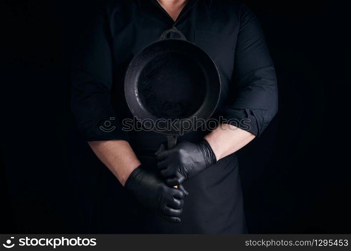 male cook in black uniform and latex gloves holds an empty round vintage black cast iron pan in front of him, low key
