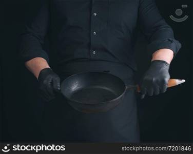 male cook in black uniform and latex gloves holds an empty round vintage black cast iron pan in front of him, low key