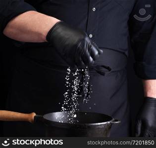 male cook in black uniform and latex gloves salt food in a black cast-iron pan, salt crystals froze in the air, low key