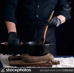 male cook in black uniform and latex gloves stirs a vintage wooden spoon food in a cast-iron pan with a handle, dark background