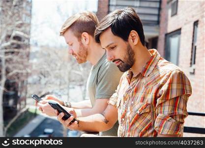 Male couple standing on balcony, looking at smartphones