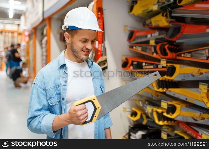 Male customer in white helmet choosing saw in hardware store. Buyer look at the goods in diy shop, shopping in building supermarket. Male customer choosing saw in hardware store