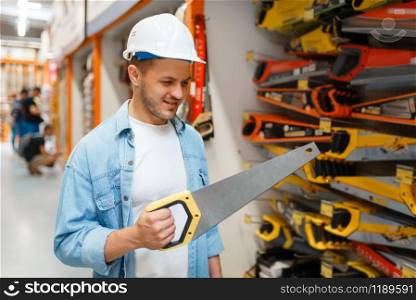 Male customer in white helmet choosing saw in hardware store. Buyer look at the goods in diy shop, shopping in building supermarket. Male customer choosing saw in hardware store