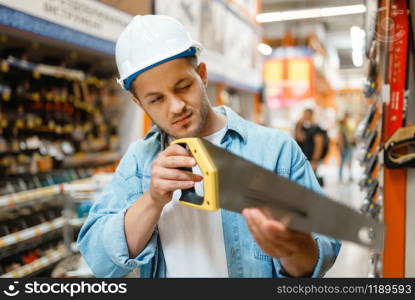 Male customer in white helmet choosing saw in hardware store. Buyer look at the goods in diy shop, shopping in building supermarket