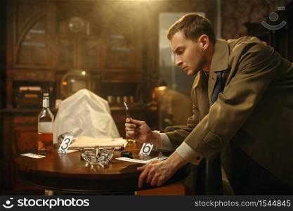 Male detective with tweezers puts the evidence in a bag at the crime scene, retro style. Criminal investigation, inspector is working on a murder, vintage room interior on background. Detective with tweezers puts the evidence in a bag