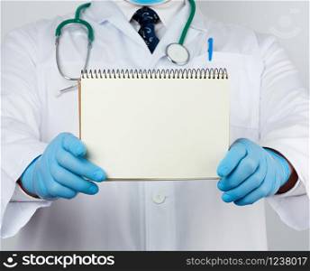 male doctor in a white coat and medical blue gloves holds a notebook with blank white sheets, place for an inscription