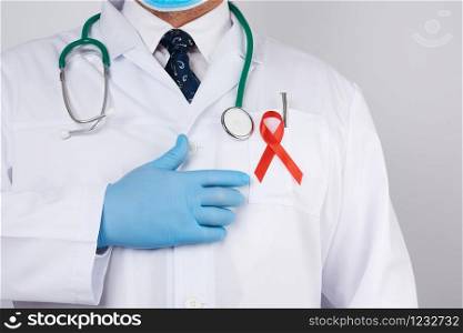 male doctor in a white coat and tie is standing on a white background, red silk ribbon is hanging on his chest, and blue sterile medical gloves are on his hands, symbol of the fight against disease AIDS and vasculitis