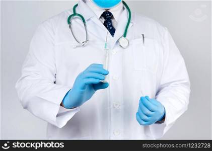 male doctor in a white coat and tie stands and holds a plastic syringe with a needle on a white background, wearing blue sterile medical gloves in his hands the concept of vaccination against diseases