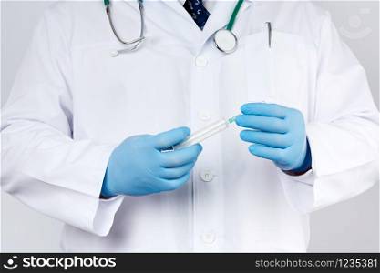 male doctor in a white coat and tie stands and holds a plastic syringe with a needle on a white background, wearing blue sterile medical gloves in his hands the concept of vaccination against diseases