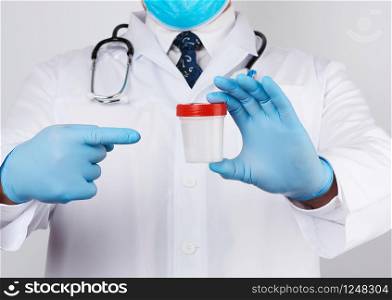 male doctor in a white coat and tie stands and holds a empty plastic container for urine specimen, wearing blue sterile medical gloves
