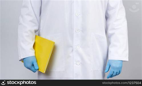 male doctor in a white coat with buttons, wearing blue sterile gloves, holding a yellow paper notebook in his hand on a white studio background