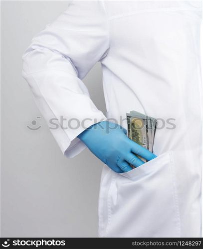 male doctor in a white medical coat and blue latex gloves puts a stack of US dollars in his coat pocket, concept of a bribe
