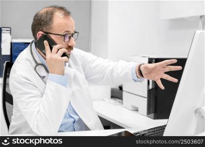 Male doctor pointing with finger on desktop computer while talking on the phone, discussing treatment with colleague. High quality photo. Male doctor pointing with finger on desktop computer while talking on the phone, discussing treatment with colleague.
