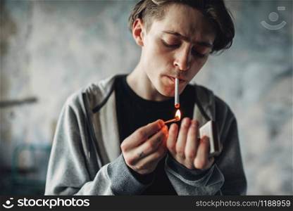 Male drug addict light a cigarette, grunge room interior on background. Smoking addiction concept. Male drug addict light a cigarette