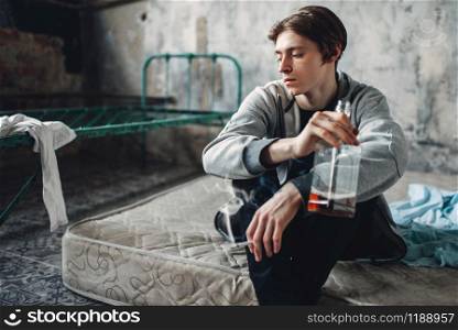 Male druggy sitting on the mattress, smoke cigarette and drinks alcohol, grunge room interior on background. Addiction concept. Male druggy smoke cigarette and drinks alcohol
