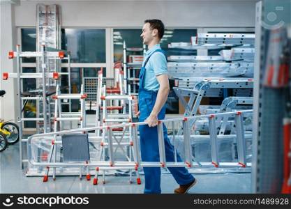 Male employee in uniform holds new aluminum stepladders in tool store. Department with ladders, choice of equipment in hardware shop, instrument supermarket. Employee holds aluminum stepladders in tool store