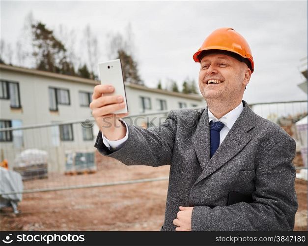 Male engineer take selfie, he wearing the suit and the hard hat, house construction site on background