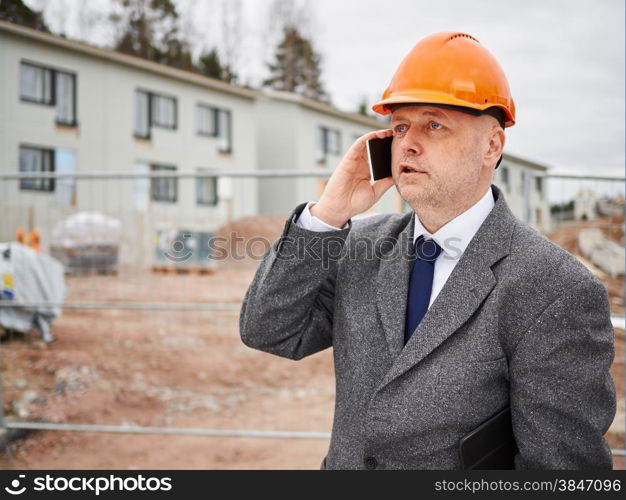 Male engineer uses cell phone, he wearing the suit and the hard hat, house construction site on background