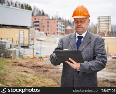 Male engineer uses tablet computer, he wearing the suit and the hard hat, house construction site on background
