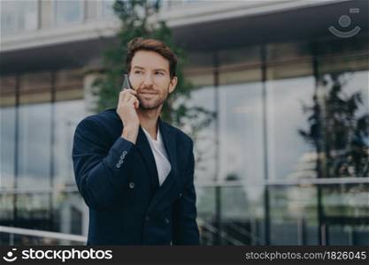 Male entrepreneur talks via phone solves urgent problems poses near business center dressed in black suit uses modern technologies for communication looks away thoughtfully. Technology work lifestyle. Male entrepreneur talks via phone solves urgent problems poses near business center