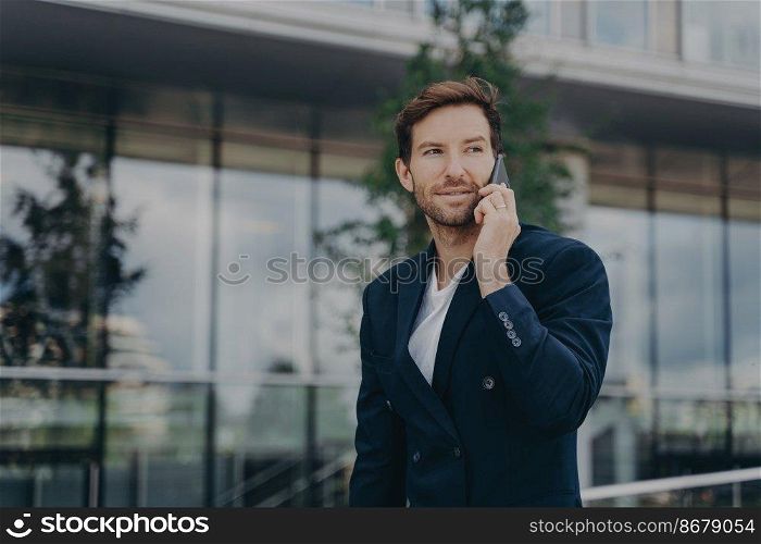 Male entrepreneur talks via phone solves urgent problems poses near business center dressed in black suit uses modern technologies for communication looks away thoughtfully. Technology work lifestyle. Male entrepreneur talks via phone solves urgent problems poses near business center