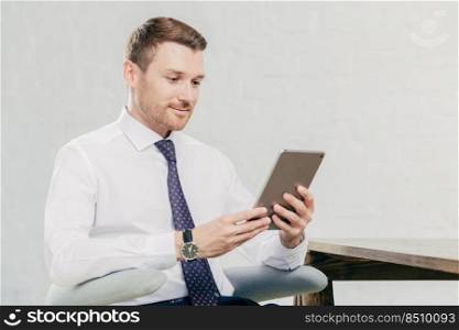 Male executive manager in white shirt and tie, holds tablet computer, searches necessary information in internet, connected to wifi in office. People, business and modern technologies concept