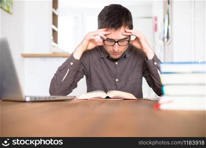 Male exhaust and unhappy student researching and learning at home, stack of books blurry
