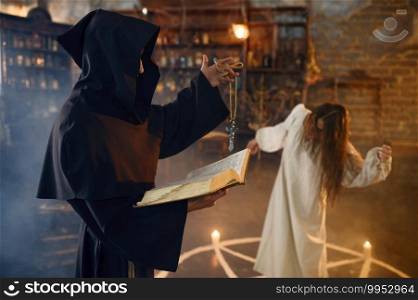 Male exorcist in black hood casting out demons from a woman. Exorcism, mystery paranormal ritual, dark religion, night horror, potions on shelf on background. Exorcist in hood casting out demons from a woman