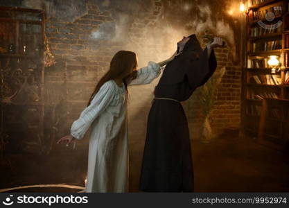 Male exorcist in black hood casting out satan from young woman. Exorcism, mystery paranormal ritual, dark religion, night horror, potions on shelf on background. Exorcist casting out satan from young woman