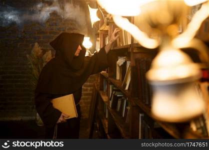 Male exorcist in black hood choosing book of spells. Exorcism, mystery paranormal ritual, dark religion, night horror, potions on shelf on background. Exorcist in black hood choosing book of spells