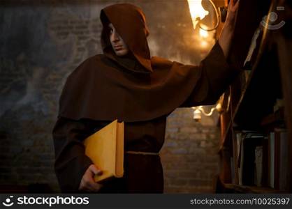 Male exorcist in black hood holding a torch. Exorcism, mystery paranormal ritual, dark religion, night horror, potions on shelf on background. Male exorcist in black hood holding a torch