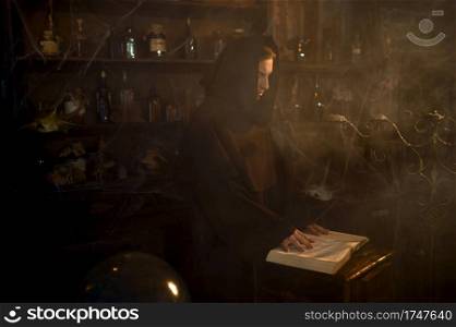 Male exorcist in black hood reads a spell. Exorcism, mystery paranormal ritual, dark religion, night horror, potions on shelf on background. Male exorcist in black hood reads a spell
