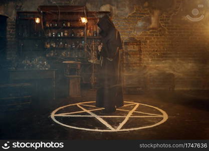 Male exorcist in black hood standing in the magic circle. Exorcism, mystery paranormal ritual, dark religion, night horror, potions on shelf on background. Male exorcist in hood standing in the magic circle