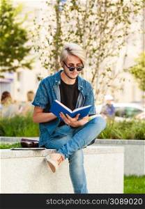 Male fashion, student concept. Guy holding and studying from notebook wearing jeans outfit and eccentric sunglasses sitting on white ledge. Hipster guy holding notebook sitting on ledge