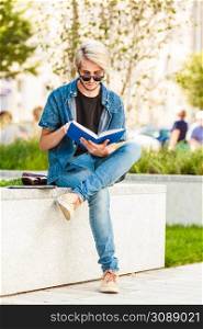 Male fashion, student concept. Guy holding and studying from notebook wearing jeans outfit and eccentric sunglasses sitting on white ledge. Hipster guy holding notebook sitting on ledge