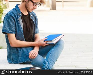 Male fashion, student concept. Guy holding notebook wearing jeans outfit and eccentric sunglasses sitting on white ledge next to modern building. Hipster guy holding notebook sitting on ledge