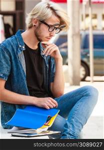 Male fashion, student concept. Guy holding notebook wearing jeans outfit and eccentric sunglasses sitting on white ledge next to modern building. Hipster guy holding notebook sitting on ledge