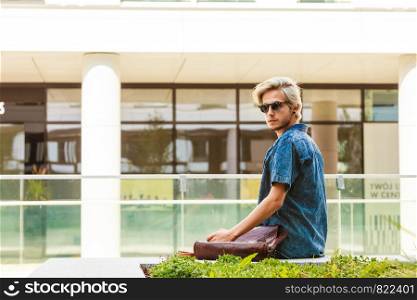 Male fashion, student concept. Guy with vintage bag wearing jeans outfit sitting on white ledge next to modern building. Guy with vintage bag sitting on ledge next to building