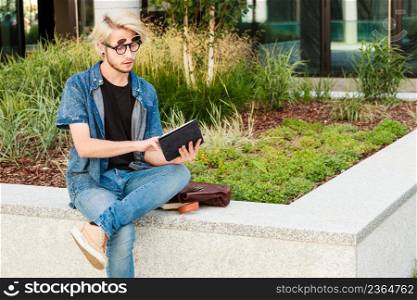 Male fashion, technology, student concept. Guy with tablet wearing jeans outfit and eccentric sunglasses sitting on white ledge. Hipster guy with tablet sitting on ledge