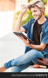 Male fashion, technology, student concept. Guy with tablet wearing jeans outfit and eccentric sunglasses sitting on white ledge. Hipster smiling guy with tablet sitting on ledge