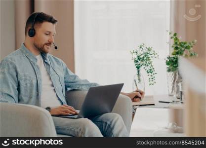 Male freelancer listens attentively tutorials how to start own business writes down information wears stereo headphones and laptop computer works online from home makes video call watches webinar