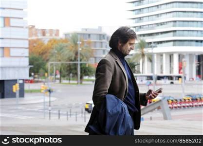 Male freelancer walking near business center holding phone, reading news, successful businessman