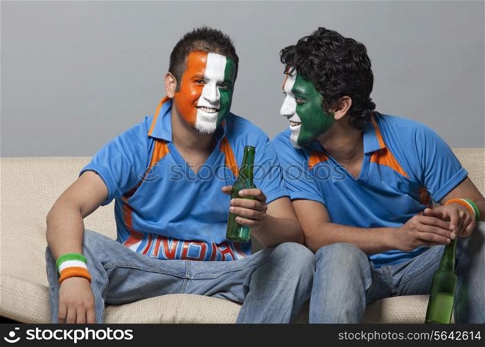 Male friends with face painted in Indian tricolor having drink while sitting on sofa at home