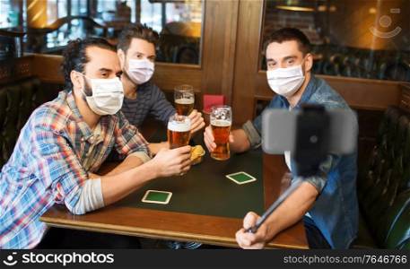 male friendship, leisure and pandemic concept - men or friends in face protective medical masks for protection from virus drinking beer and taking picture with smartphone selfie stick at bar or pub. men in masks take selfie and drink beer at bar
