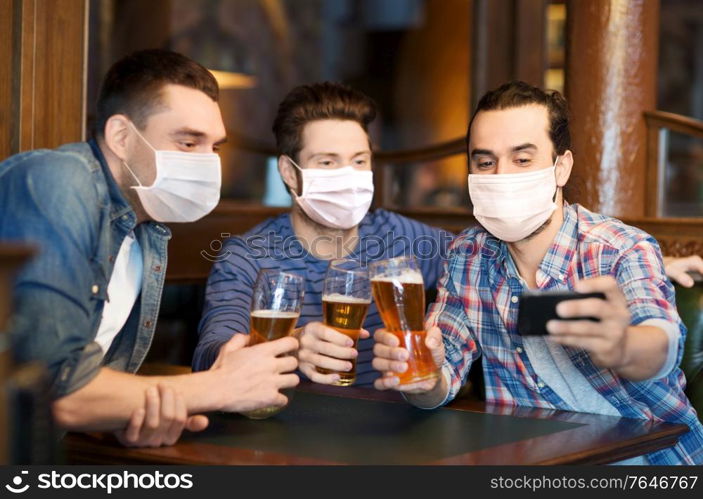 male friendship, leisure and pandemic concept - men or friends in face protective medical masks for protection from virus drinking beer and taking selfie with smartphone at bar or pub. men in masks take selfie and drink beer at bar