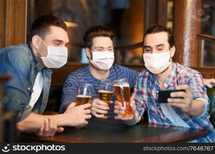 male friendship, leisure and pandemic concept - men or friends in face protective medical masks for protection from virus drinking beer and taking selfie with smartphone at bar or pub. men in masks take selfie and drink beer at bar