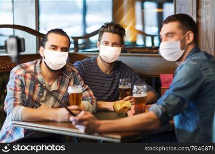 male friendship, leisure and pandemic concept - men or friends in face protective medical masks for protection from virus drinking beer and taking picture with smartphone selfie stick at bar or pub. men in masks take selfie and drink beer at bar