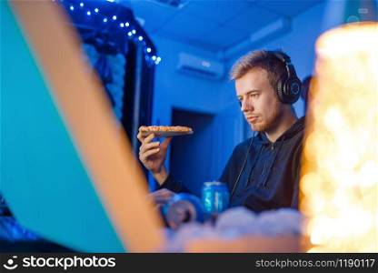 Male gamer eating pizza at his workplace with laptop and desktop PC, gaming lifestyle, night tournament. Computer games player in his room with neon light, streamer. Male gamer eating pizza, night tournament